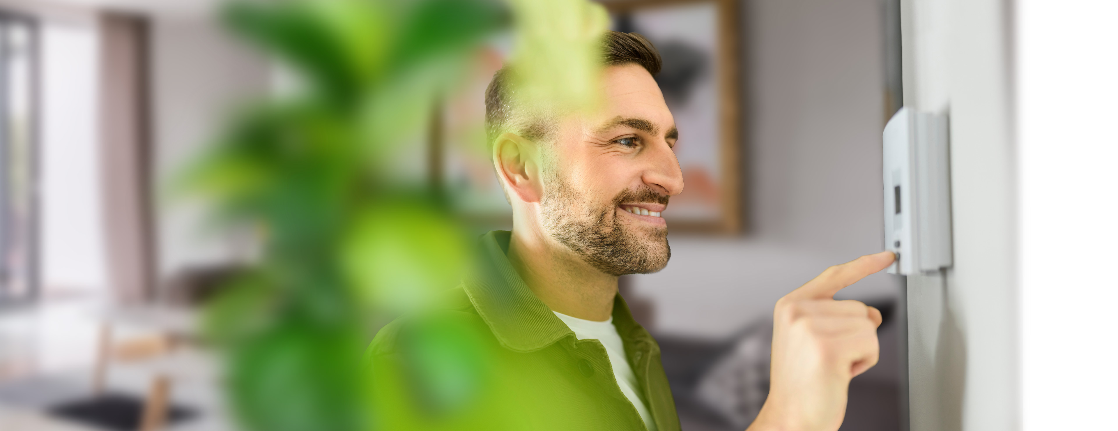 Junger Mann steht lächelnd vor seinem Thermostat und stellt es ein.