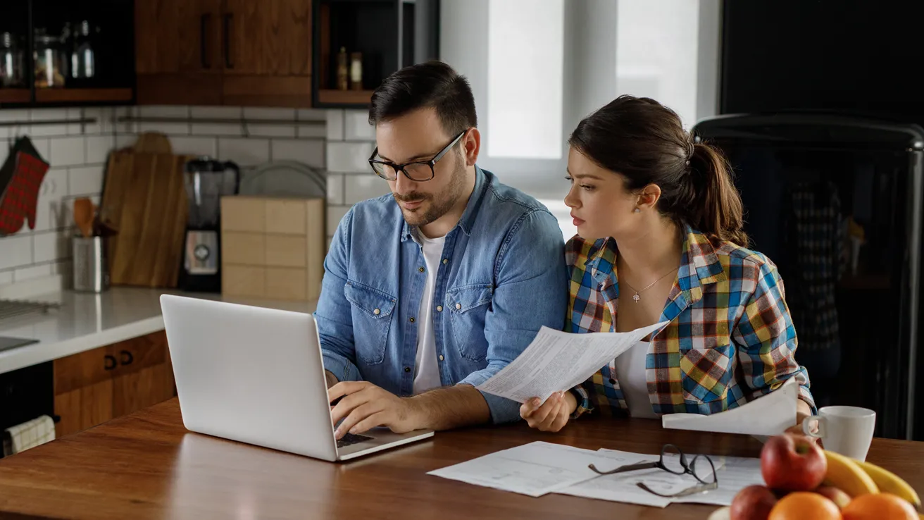 Ehepaar sitzt in Küche vor dem Laptop und schließt einen Vertrag ab.