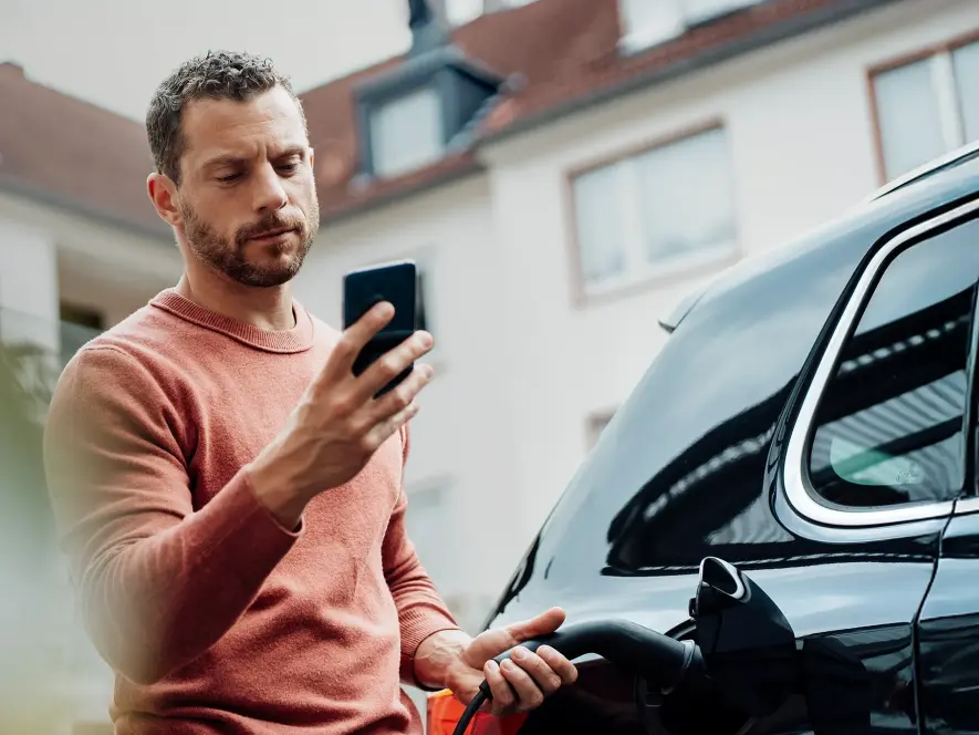 Ein Mann lädt sein schwarzes Elektroauto und schaut dabei auf sein Handy.