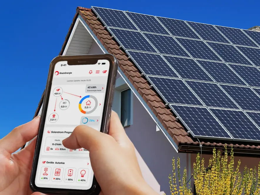 Eine Hand hält ein Smartphone mit geöffenter RheinSmart-App vor einem Einfamilienhaus mit Solaranlage auf dem Dach