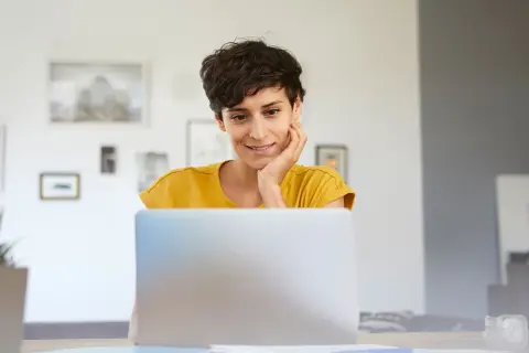 Frau mit dunklen Haaren sitzt am Tisch und schaut auf einen Laptop-Bildschirm