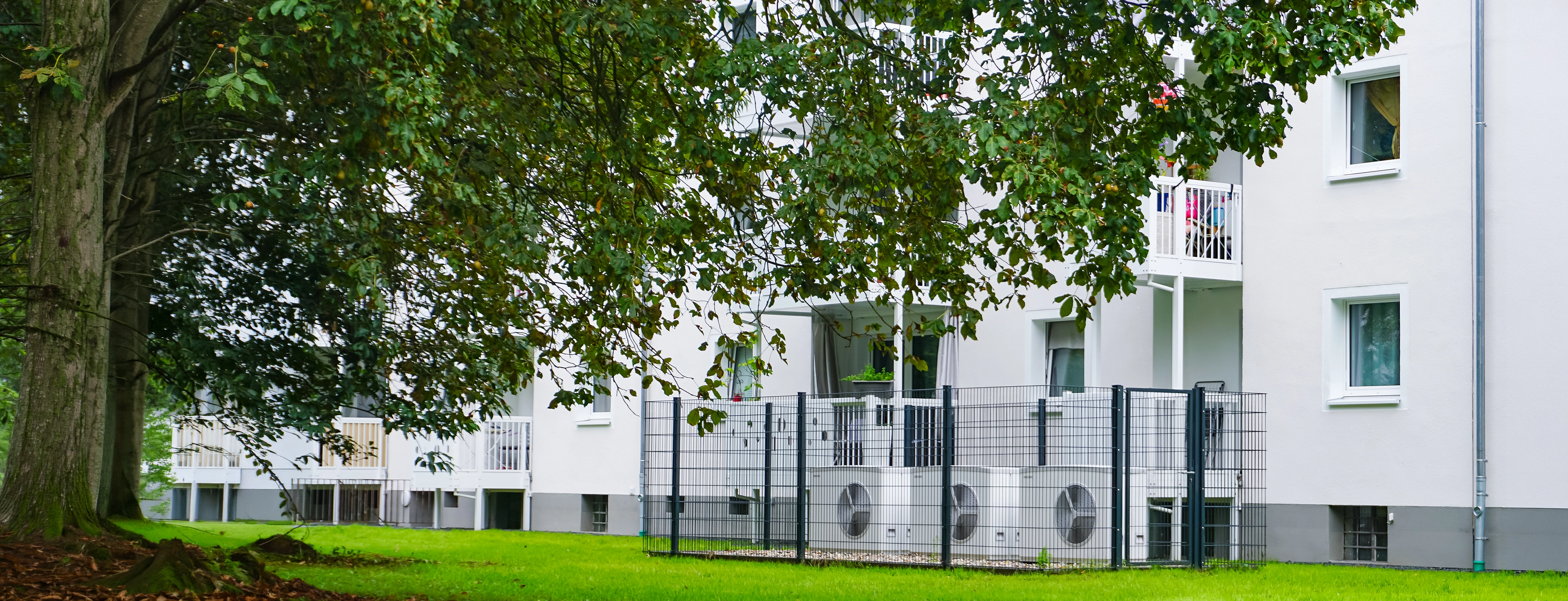 Eingezäunte Wärmepumpen vor einem Mehrfamilienhaus.