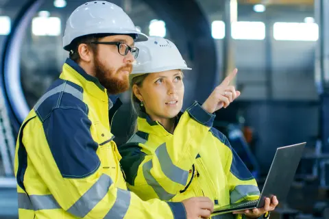 Mann und Frau in Sicherheitskleidung und Helm in einer Produktionshalle.