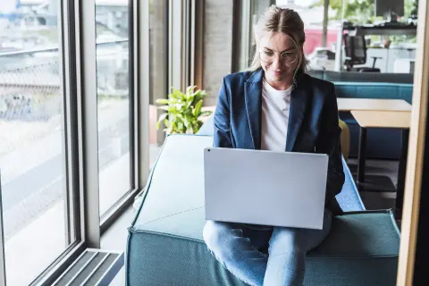 Eine junge Frau sitzt auf der Sofalehne und arbeitet am Laptop.