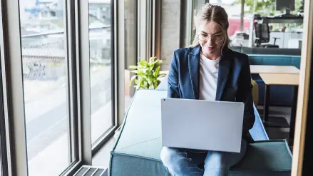 Junge Frau sitzt auf Sofalehne und arbeitet am Laptop.