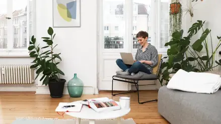 Frau sitzt mit Laptop auf Schoß im Schneidersitz in einem Wohnzimmer. 