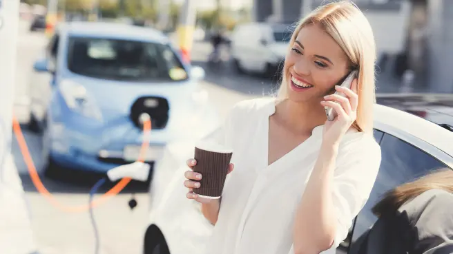 Frau lädt ihr E-Auto an einer Ladestation und telefoniert währenddessen