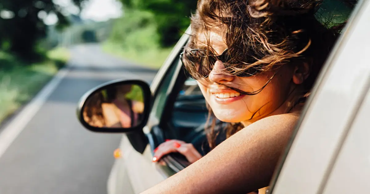 Eine Frau mit Sonnenbrille schaut aus dem Fahrerfenster eines Autos auf einer Landstraße