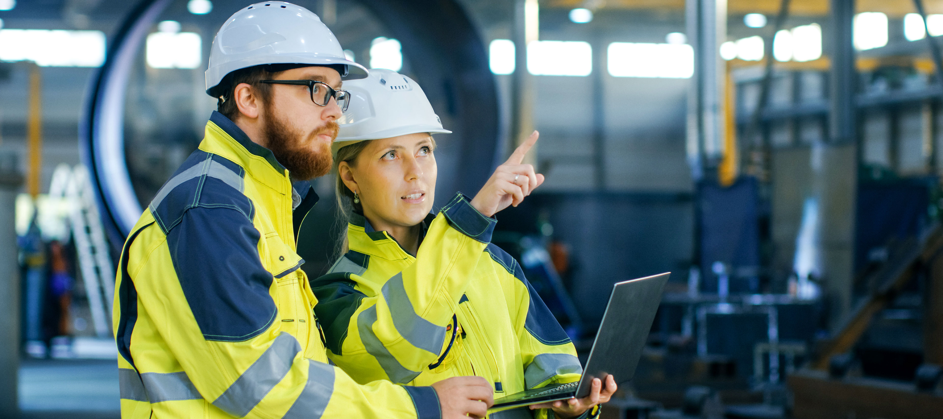 Mann und Frau in Sicherheitskleidung und Helm in einer Produktionshalle.